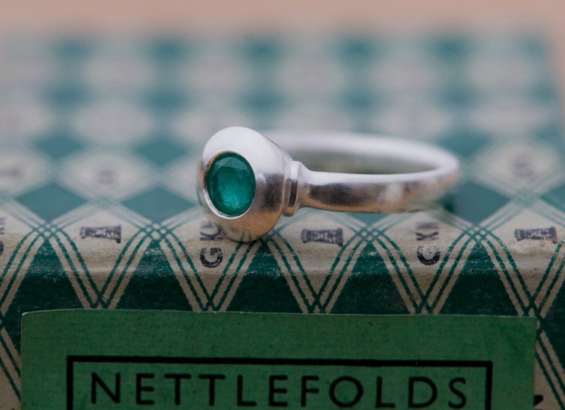 May include: A silver ring with a green gemstone set in a round bezel. The ring is sitting on a green and white patterned box with the word "NETTLEFOLDS" printed on the front.