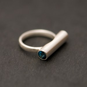 Pu&ograve; includere: Un anello d'argento con una pietra preziosa blu incastonata al centro di una fascia cilindrica.