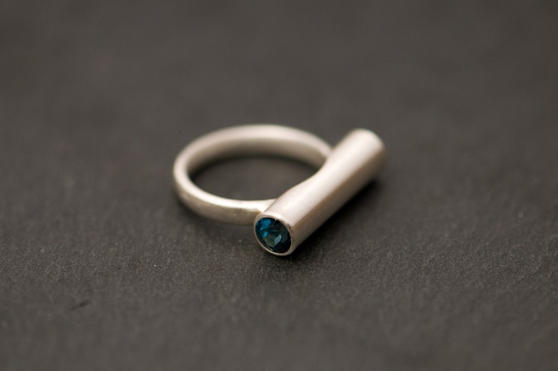 Pu&ograve; includere: Un anello d'argento con una pietra preziosa blu incastonata al centro di una fascia cilindrica.