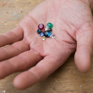 May include: A hand holding a variety of colorful gemstones, including red, blue, green, and yellow.