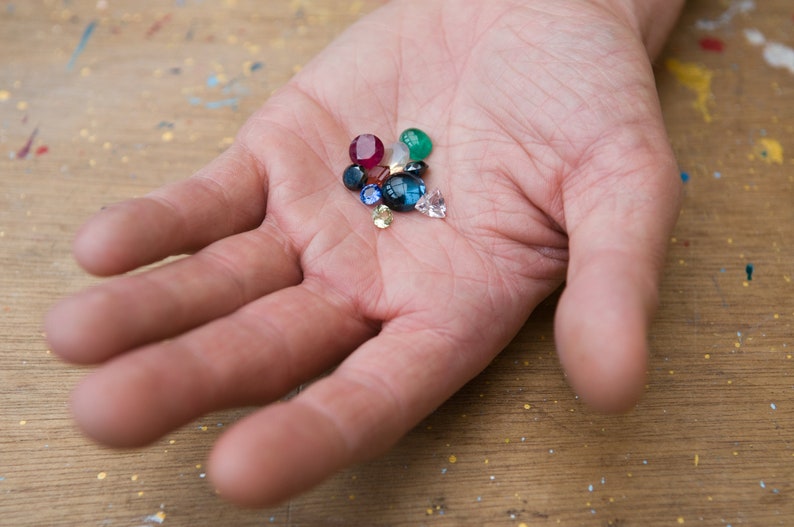 May include: A hand holding a variety of colorful gemstones, including red, blue, green, and yellow.