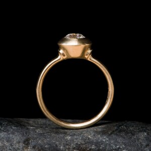 May include: A gold ring with a bezel setting featuring a round, clear gemstone. The ring is on a dark grey rock.