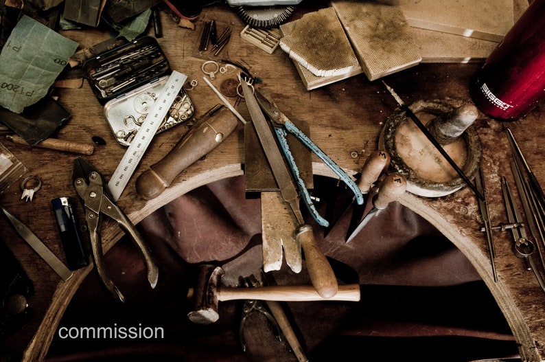 May include: A wooden workbench with various tools and supplies for jewelry making, including pliers, a hammer, a ruler, a file, and a small box of tools. The workbench is covered in a brown leather surface.