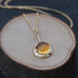 May include: A gold necklace with a pendant featuring a round, amber-colored gemstone set in a gold bezel. The pendant hangs from a delicate gold chain.