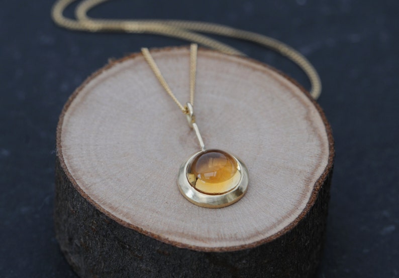 May include: A gold necklace with a pendant featuring a round, amber-colored gemstone set in a gold bezel. The pendant hangs from a delicate gold chain.