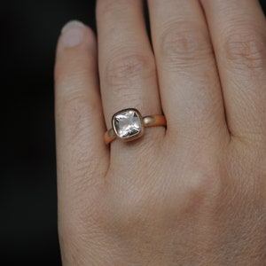 May include: A gold ring with a square-shaped, clear gemstone. The ring is on a person's finger.