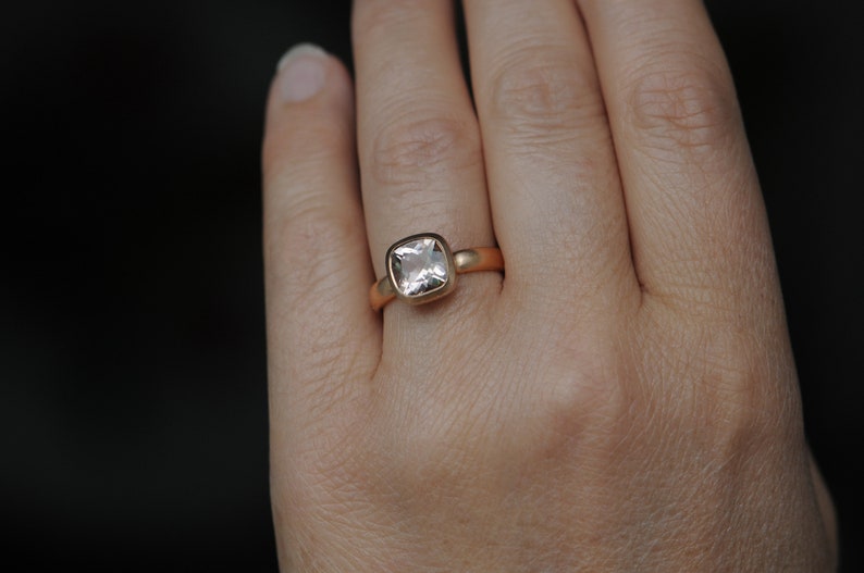 May include: A gold ring with a square-shaped, clear gemstone. The ring is on a person's finger.