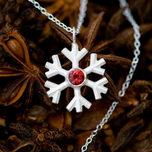 Könnte beinhalten: Eine silberne Schneeflocken-Anhänger-Kette mit einem roten Edelstein in der Mitte. Die Kette liegt auf einem Bett aus braunem Sternanis.