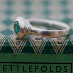May include: A silver ring with a green gemstone set in a bezel setting. The ring is on a green and white patterned surface.