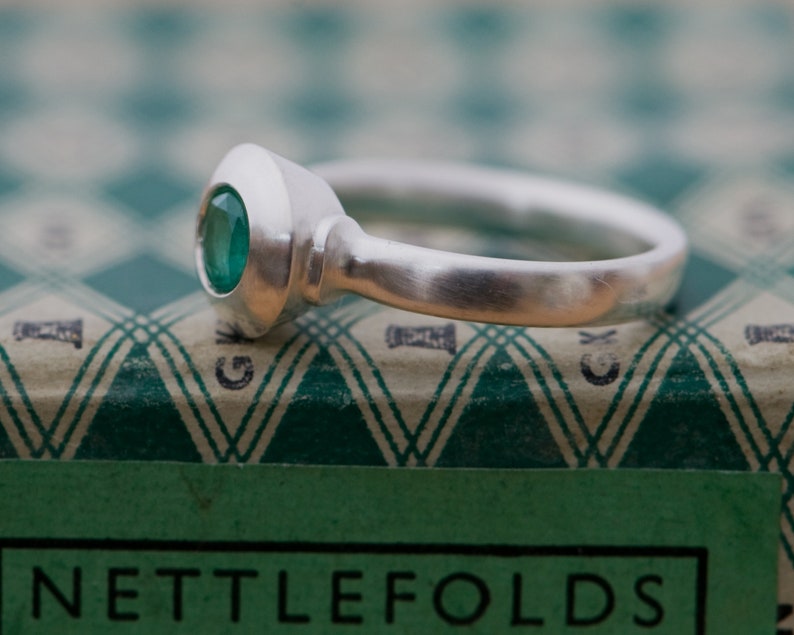May include: A silver ring with a green gemstone set in a bezel setting. The ring is on a green and white patterned surface.