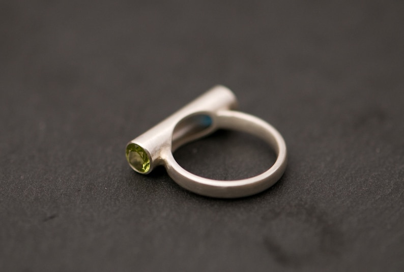 Pu&ograve; includere: Un anello d'argento con una pietra preziosa verde incastonata in una fascia cilindrica.