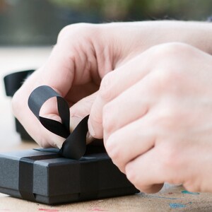 May include: A black gift box with a black ribbon tied in a bow. The ribbon is being held by a hand.