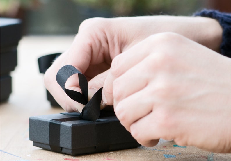 May include: A black gift box with a black ribbon tied in a bow. The ribbon is being held by a hand.