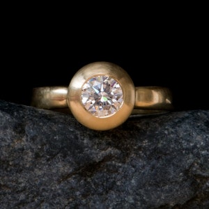 May include: A gold ring with a round bezel setting featuring a clear diamond. The ring is resting on a dark grey rock.