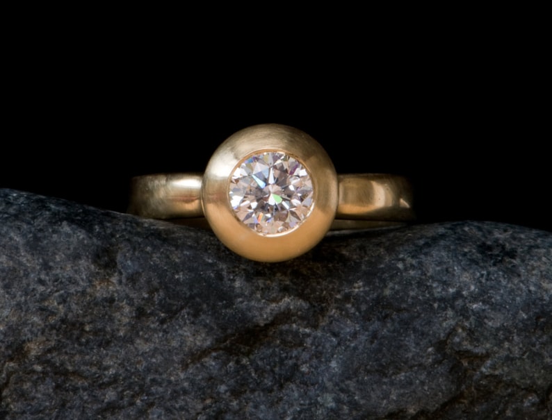 May include: A gold ring with a round bezel setting featuring a clear diamond. The ring is resting on a dark grey rock.