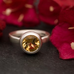 May include: A silver ring with a round, yellow gemstone set in a bezel setting. The ring is on a dark gray surface with red flower petals in the background.