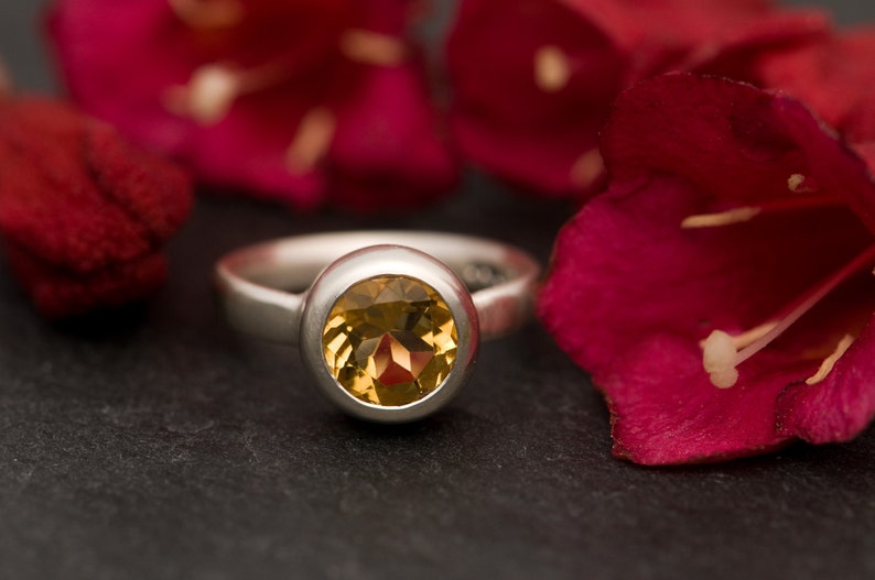 May include: A silver ring with a round, yellow gemstone set in a bezel setting. The ring is on a dark gray surface with red flower petals in the background.