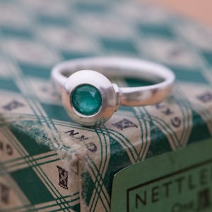 May include: A silver ring with a round, bezel-set emerald green gemstone. The ring is sitting on a green and white patterned box.