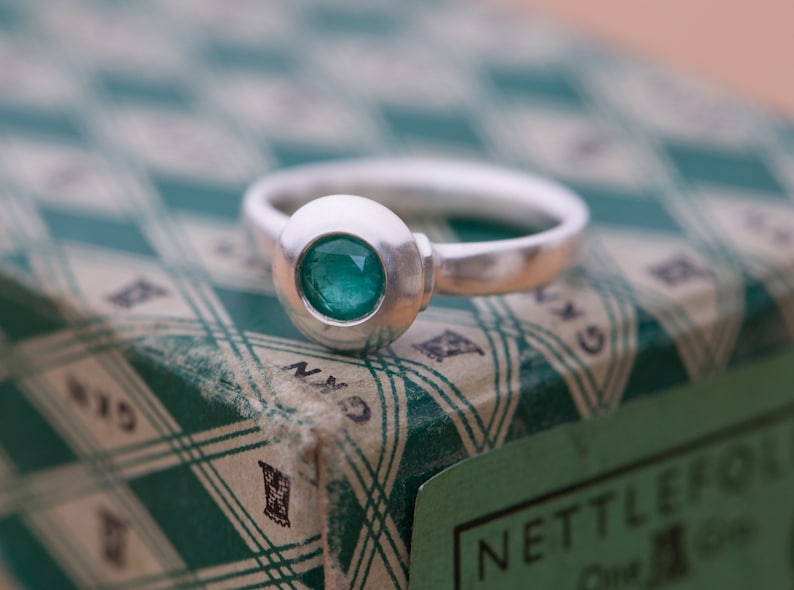 May include: A silver ring with a round, bezel-set emerald green gemstone. The ring is sitting on a green and white patterned box.