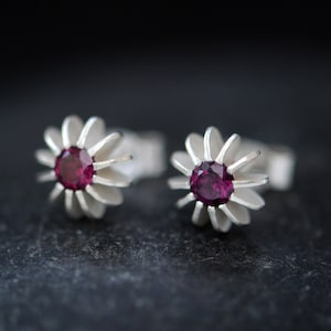 May include: A pair of silver earrings with a flower design and a small, dark purple gemstone in the center of each flower.