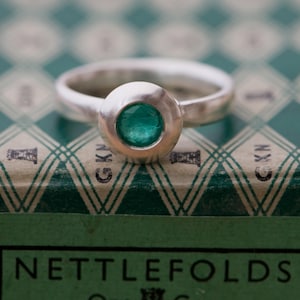 May include: A silver ring with a round bezel set with a green gemstone. The ring is sitting on a green and white patterned box with the text "Nettlefolds" printed on the front.