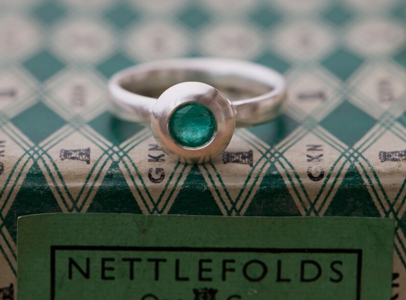 May include: A silver ring with a round bezel set with a green gemstone. The ring is sitting on a green and white patterned box with the text "Nettlefolds" printed on the front.