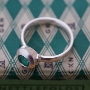 May include: A silver ring with a round, bezel-set emerald green gemstone. The ring is on a green and white patterned surface.