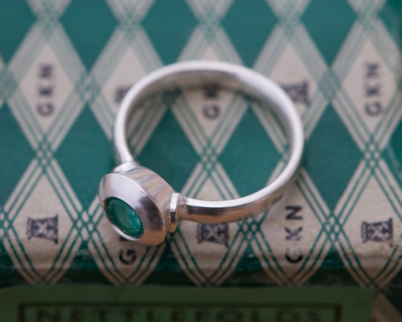 May include: A silver ring with a round, bezel-set emerald green gemstone. The ring is on a green and white patterned surface.
