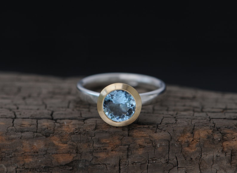 Puede incluir: Un anillo de plata con una piedra preciosa azul redonda engastada en un engaste de oro.