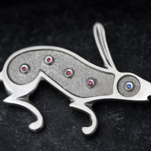 May include: A silver rabbit brooch featuring red and blue gemstones. The rabbit is depicted in a running pose with its ears raised. The brooch has a textured surface.