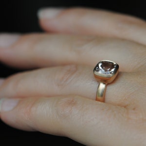 May include: A gold ring with a light pink square-shaped gemstone. The ring is on a person's finger.