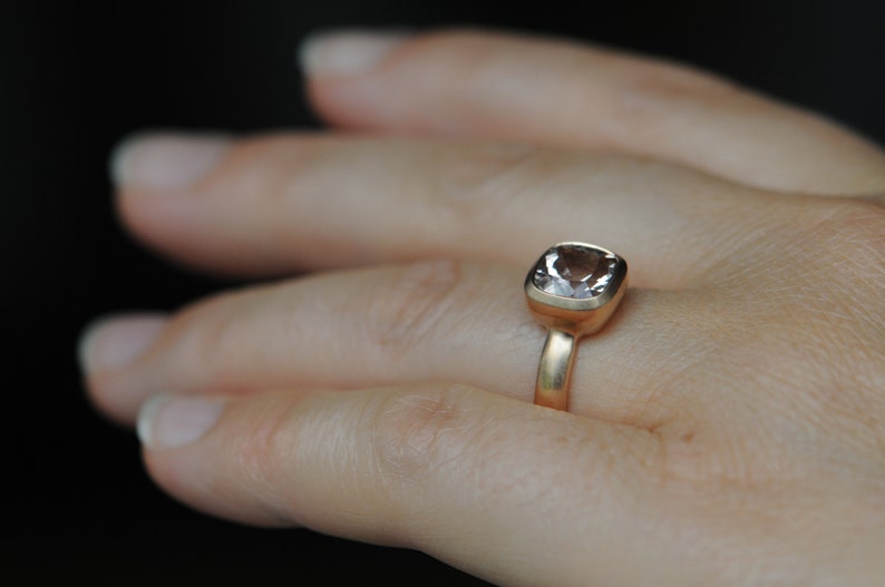May include: A gold ring with a light pink square-shaped gemstone. The ring is on a person's finger.
