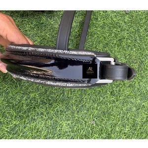 May include: A close-up of a black horse bridle with a glossy finish. The bridle features a row of sparkling rhinestones along the top and bottom edges. The brand logo "AR ALISH" is visible on the bridle.