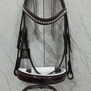 May include: A brown leather horse bridle with a white padded noseband and a browband adorned with sparkling rhinestones. The bridle includes a matching throat latch and a separate, adjustable noseband. The brand name "ALISH" is visible.