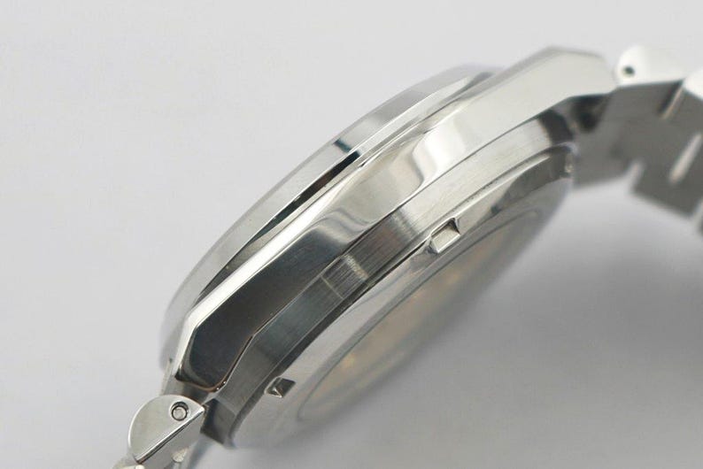 May include: Close-up of a silver wristwatch with a metallic band. The watch case has a polished finish and a sleek, modern design. The watch band is made of interlocking metal links.