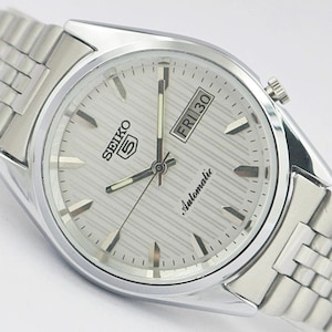 Peut inclure: Une montre-bracelet automatique Seiko 5 argentée avec un cadran blanc texturé, des index bâtons argentés et un guichet jour-date à trois heures. La montre est dotée d'un bracelet à maillons en acier inoxydable et d'un boîtier argenté poli.
