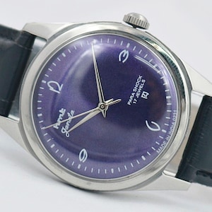 May include: A vintage wristwatch with a silver-toned case and a deep purple dial. The watch features silver hands and hour markers, with the text "Para Shock 17 Jewels" and "Ame Janata" on the face. It has a black leather band.