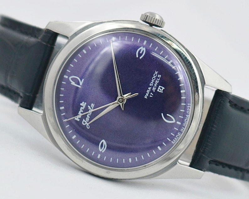 May include: A vintage wristwatch with a silver-toned case and a deep purple dial. The watch features silver hands and hour markers, with the text "Para Shock 17 Jewels" and "Ame Janata" on the face. It has a black leather band.