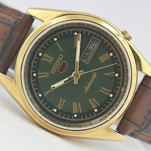 May include: A gold-tone Seiko 5 automatic wristwatch featuring a dark green dial with Roman numeral hour markers, gold hands, and a day-date window at the three o'clock position. The watch is attached to a brown leather strap with a textured finish.