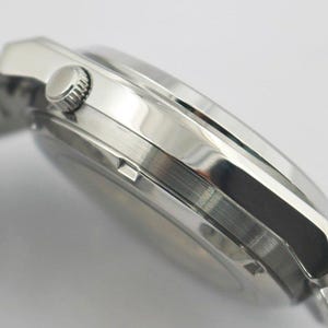 May include: Close-up of a silver wristwatch with a stainless steel band. The watch has a round face and a textured crown. The watch band is made of interlocking metal links. The watch is angled to show the side profile.