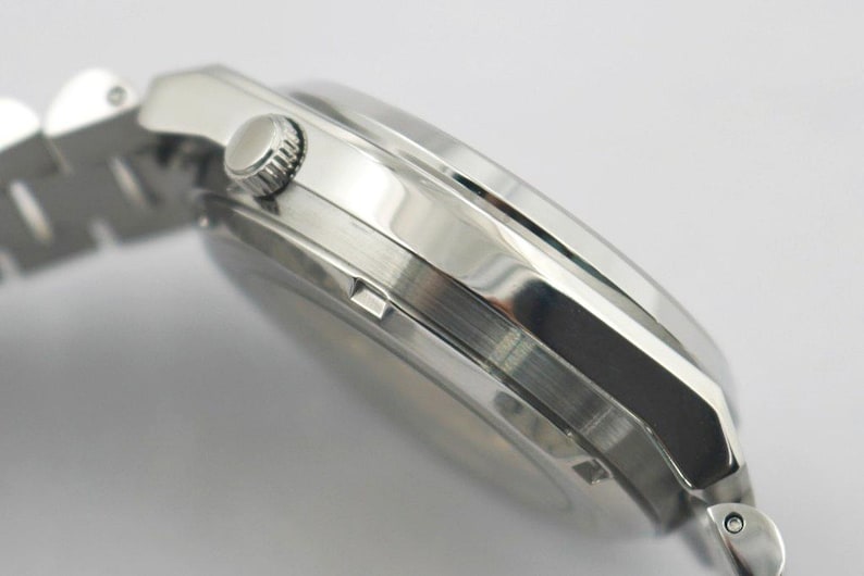 May include: Close-up of a silver wristwatch with a stainless steel band. The watch has a round face and a textured crown. The watch band is made of interlocking metal links. The watch is angled to show the side profile.
