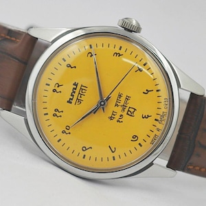 Könnte beinhalten: Eine silberfarbene Armbanduhr mit gelbem Zifferblatt und braunem Lederarmband. Das Zifferblatt hat schwarze Ziffern und Text in einer Fremdsprache. Das Armband hat ein Krokodilmuster.