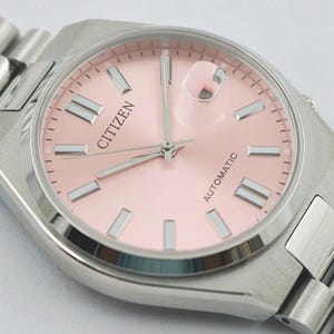 May include: A Citizen automatic wristwatch with a light pink dial and silver-tone hands and markers. The watch has a stainless steel case and a matching link bracelet. The word "CITIZEN" is printed on the dial.