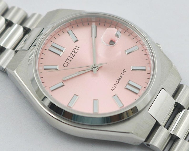May include: A Citizen automatic wristwatch with a light pink dial and silver-tone hands and markers. The watch has a stainless steel case and a matching link bracelet. The word "CITIZEN" is printed on the dial.