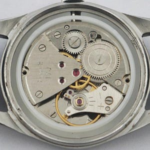 May include: Close-up of a vintage wristwatch with the back removed, revealing the intricate mechanical movement. The watch features silver-colored gears, ruby jewels, and a black leather strap.