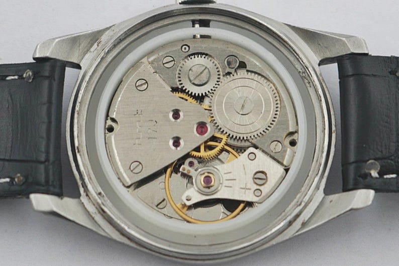 May include: Close-up of a vintage wristwatch with the back removed, revealing the intricate mechanical movement. The watch features silver-colored gears, ruby jewels, and a black leather strap.
