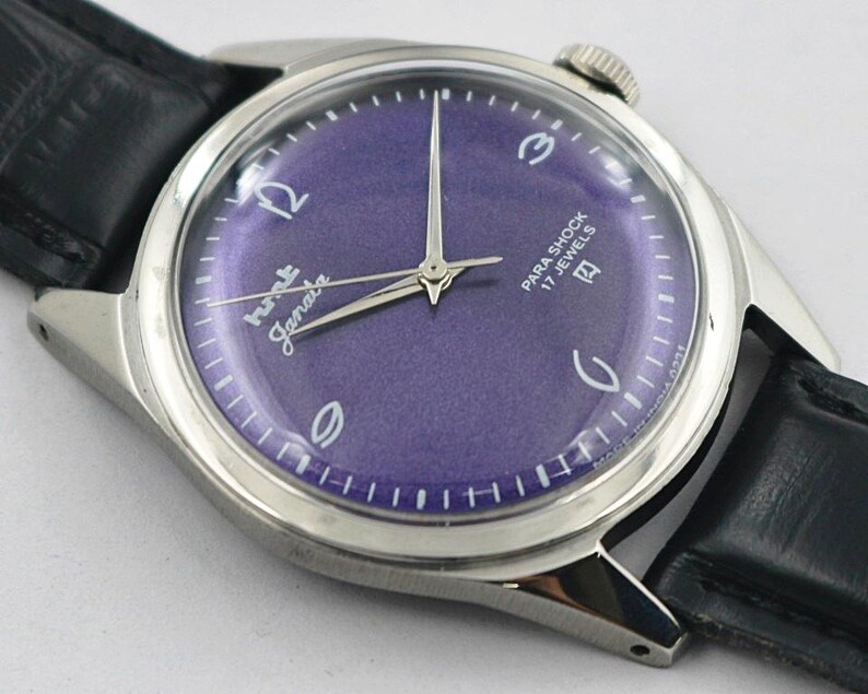 May include: A vintage wristwatch with a purple dial and silver hands. The watch face has white numerals and the words "Para Shock 17 Jewels". The watch has a silver case and a black leather band.