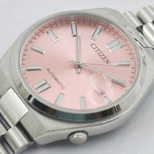 May include: A Citizen automatic wristwatch with a light pink dial and silver-tone hands and markers. The watch has a stainless steel case and a matching bracelet. The word "CITIZEN" is printed on the dial.
