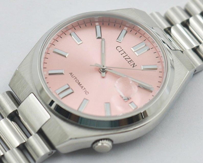 May include: A Citizen automatic wristwatch with a light pink dial and silver-tone hands and markers. The watch has a stainless steel case and a matching bracelet. The word "CITIZEN" is printed on the dial.