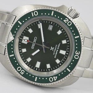Può includere: Un orologio subacqueo automatico Seiko in acciaio inossidabile con quadrante e lunetta verde scuro. L'orologio presenta indici, lancette e un datario bianchi a ore 3. Il cinturino è in acciaio inossidabile.